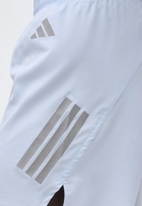 Λευκά αθλητικά σορτς με τσέπη στο αριστερό μηρό, η οποία διαθέτει τρεις κάθετες ανακλαστικές ρίγες και λογότυπο Adidas πάνω από την τσέπη.