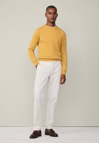 Žlutý pletený svetr s kulatým výstřihem, doplněný bílými kalhotami. Hnědé mokasíny s třásněmi dotvářejí outfit. Neutrální pozadí.