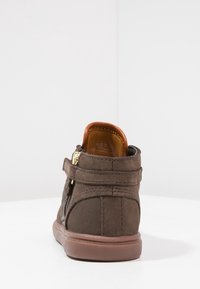 Zapato de tobillo de gamuza marrón con una textura suave, que presenta una cremallera lateral, cierre de correa y una suela de goma marrón.