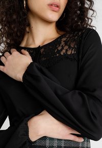 Top nero con scollatura in pizzo, maniche lunghe e a sbuffo, e corpo aderente. Tessuto morbido con dettagli in pizzo intorno al colletto.