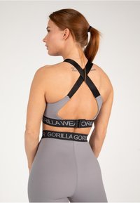 Sujeción deportiva gris con un diseño de corte, que presenta tirantes en forma de X negros y una cinturilla de marca en contraste. Textura de tela suave.