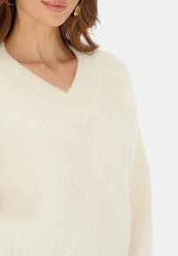 Sweter w kształcie litery V wykonany z miękkiego, kremowego materiału. Charakteryzuje się luźnym krojem oraz prążkowanym wykończeniem dekoltu. Tekstura wydaje się gładka i puszysta.