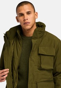 Parka de color verde oliva con un cuello alto, cremallera frontal y múltiples bolsillos en el pecho. Llevada sobre un suéter de textura verde oscuro.