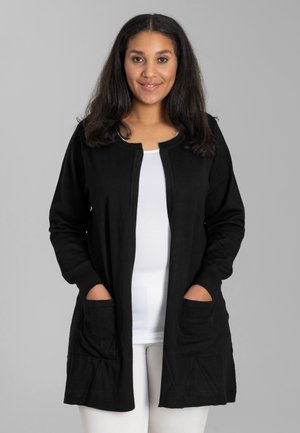 Cardigan nero realizzato in tessuto morbido, con maniche lunghe, scollo rotondo, due tasche frontali e una vestibilità comoda, abbinato a una maglietta bianca.