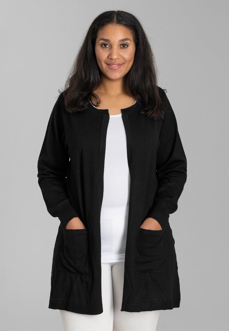Schwarze Strickjacke aus weichem Stoff, mit langen Ärmeln, rundem Ausschnitt, zwei vorderen Taschen und lässiger Passform, kombiniert mit einem weißen Oberteil.