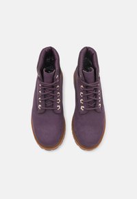 Botas de tobillo de ante morado con suelas de goma marrón gruesas, con ojales de metal y cordones a juego, forro interior suave y cuello acolchado.