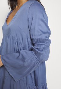 Žena v volných modrých šatech s dlouhým rukávem, které mají na rukávech a pod prsy nařasené detaily.