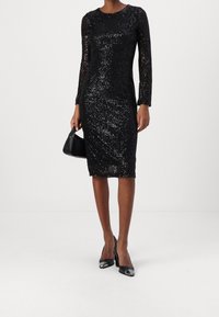 Robe noire à paillettes avec des manches longues et un col rond, ajustée sur l'ensemble, associée à des escarpins noirs et un petit sac à main.