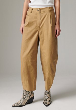 Personne portant un pantalon ample beige coupé aux chevilles et des bottes à talons hauts pointues avec motif peau de serpent sur un fond uni.