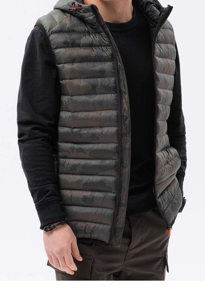 Gepolsterte Weste mit einem Tarnmuster in Grautönen und Grün, mit Reißverschluss und einem schwarzen Langarmshirt darunter.