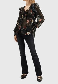 Femme portant un chemisier noir transparent à motifs floraux avec des volants, un pantalon noir ajusté et des escarpins noirs pointus avec des boucles dorées, debout devant un fond blanc.