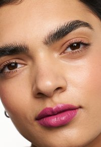 Gros plan sur un visage avec des sourcils foncés bien marqués, une peau lisse et des lèvres brillantes rose vif. Le teint est chaud avec une texture subtile.