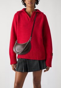 Maglione rosso lavorato a maglia con scollo a V e polsini a coste, abbinato a una gonna plissettata a quadri neri. Borsa a spalla in pelle marrone con catena argentata.