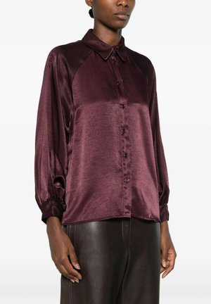 Donna che indossa una camicetta di raso viola scuro lucida con maniche lunghe, bottoni e colletto e pantaloni in pelle marrone scuro.