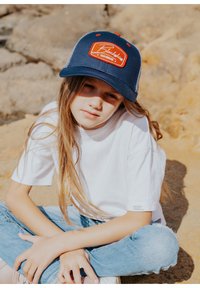 Navy cap with red label reading "Blackbies Trademark," worn with a white T-shirt and light blue jeans, against a rocky background.