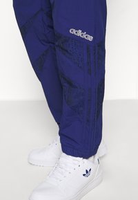 Pantaloni della tuta Adidas blu navy con dettagli neri a motivo sui lati, dotati di una vita elasticizzata e orli a coste; abbinati a sneaker bianche.