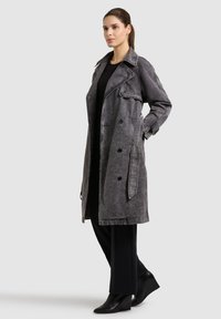 Pruunide juustega naine, kellel on seljas pikk tuhmmust mustast trentšmantlist, mustad püksid ja mustad kontsaga saabad, seisab käed taskutes.