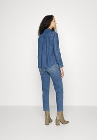 Casaco de ganga e calças em azul médio, com um corte descontraído. Botas de salto alto, em verde-oliva, com cano até ao tornozelo. Textura suave, design simples e detalhes em botão.