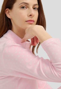 Top de forro polar rosa claro con patrón de corazones blancos, que presenta una textura suave y un corte relajado. Cuello abotonado y mangas largas.