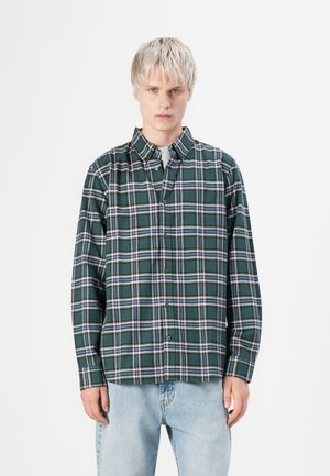 Jeune personne avec des cheveux blonds courts portant une chemise à carreaux verte et bleue et un jean bleu clair, se tenant devant un fond blanc.