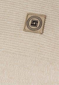 Beige stickat tyg med en texturerad väv, som har en fyrkantig patch med en svart geometrisk logotyp i en cirkulär kontur.