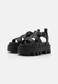 Black platform sandals with a thick, jagged sole, featuring crisscross leather straps and a side buckle for adjustable fit.