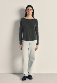 Nainen seisoo vaaleaa seinää vasten, yllä tummanharmaa pitkähihaisen paita, valkoiset väljät farkut ja mustat loaferit, katse suoraan eteenpäin.