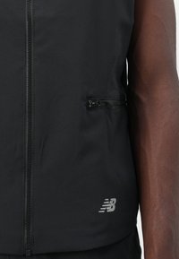 Crni sportski prsluk s punim prednjim zatvaračem, bočnim džepovima sa zatvaračem i logotipom New Balance. Napravljen od lagane, teksturirane tkanine.