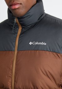 Columbia pufferjack met een hoge kraag; de bovenhelft is zwart en de onderhelft bruin. Heeft een rits en het merkteken op de borst.