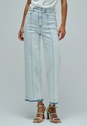 Mujer con vaqueros cortos de tiro alto, lavado claro, con dobladillos sin rematar y sandalias beige de tacón alto con tiras sobre un fondo liso.