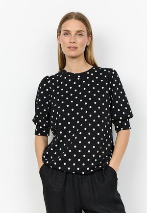 Woman with straight blonde hair wearing a black short-sleeve blouse with white polka dots and black pants, hands in pockets.