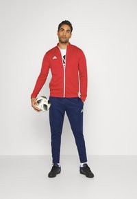 Czerwona sportowa kurtka z zamkiem błyskawicznym, z białymi akcentami i logo Adidas; granatowe spodnie sportowe; trzyma czarno-biały wzorzysty piłkę nożną.