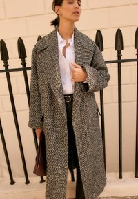 Manteau surdimensionné en cheque gris avec grands revers, devant ouvert et poches latérales. Associé à une chemise blanche et un pantalon sombre.