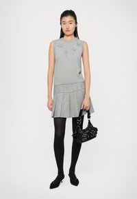 Maglione senza maniche grigio con dettagli floreali, abbinato a una gonna plissettata grigia. La modella tiene in mano una borsa nera con hardware argentato e indossa collant neri.