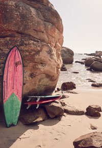 Surfboards leaning against a large rocky formation. One board is pink and green; others are red and green. Sand and water in the background.