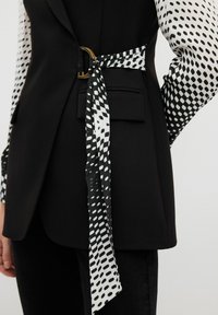 Blaser noir avec une cravate noire et blanche à motifs à la taille, présentant un accent en anneau doré et des manches texturées avec un motif à pois.