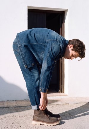 Un homme en veste et pantalon en jean se penche pour ajuster le revers de son pantalon au-dessus de ses bottes marron à lacets, devant une porte.