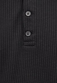 Μαύρο ύφασμα πλεκτό σε σχέδιο βάφλας με δύο μαύρα κουμπιά σε πλάκα, που δείχνει υφή και λεπτομέρειες ραφής.