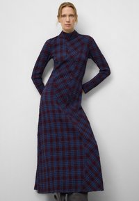 Langärmliges Kleid im dunklen burgunder und blauen Karo-Muster, mit hohem Kragen und schrägen Nahtdetails, aus strukturiertem Stoff gefertigt.