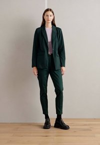 Temno zelena fasonirana blazer in usklajene slim-fit hlače, v kombinaciji s svetlo rožnato majico in črnimi čevlji, postavljeni na nevtralno ozadje.