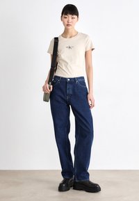 Bežové bavlnené tričko s krátkymi rukávmi a malým logom, skombinované s tmavomodrými nohavicami s vysokým pásom. Modelka drží zelenú crossbody tašku.