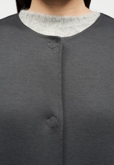 Γυναίκα που φοράει γκρι υφασμάτινο σακάκι πάνω από ανοιχτό γκρι πλεκτό πουλόβερ, δείχνοντας τα δύο εξάγωνα ραμμένα κουμπώματα του σακακιού.