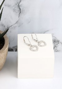 Silver hjärtformade örhängen med klara kristallaccenter, med en låsning av typ leverback, visade på en vit låda. Texturerad bakgrund.