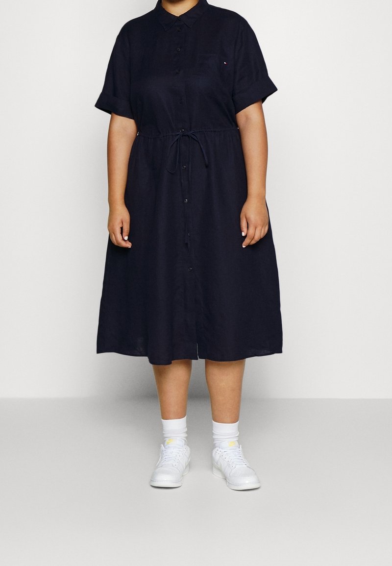 Robe chemise marine à manches courtes, devant boutonné, taille cintrée et jupe longueur genou. Portée avec des baskets blanches et des chaussettes.