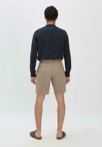 Herenoutfit bestaande uit een donker, lang mouwenshirt, beige shorts en bruine instap sandalen, gezien van de achterkant. De shorts hebben achterzakken.