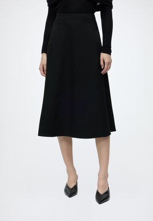 Vrouw met een zwarte top met lange mouwen, een zwarte midi A-lijn rok en zwarte puntige hakken tegen een effen witte achtergrond.