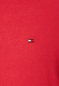 Tissu tricoté rouge avec un petit logo brodé comportant des rayures horizontales en blanc, rouge et noir en haut à gauche.