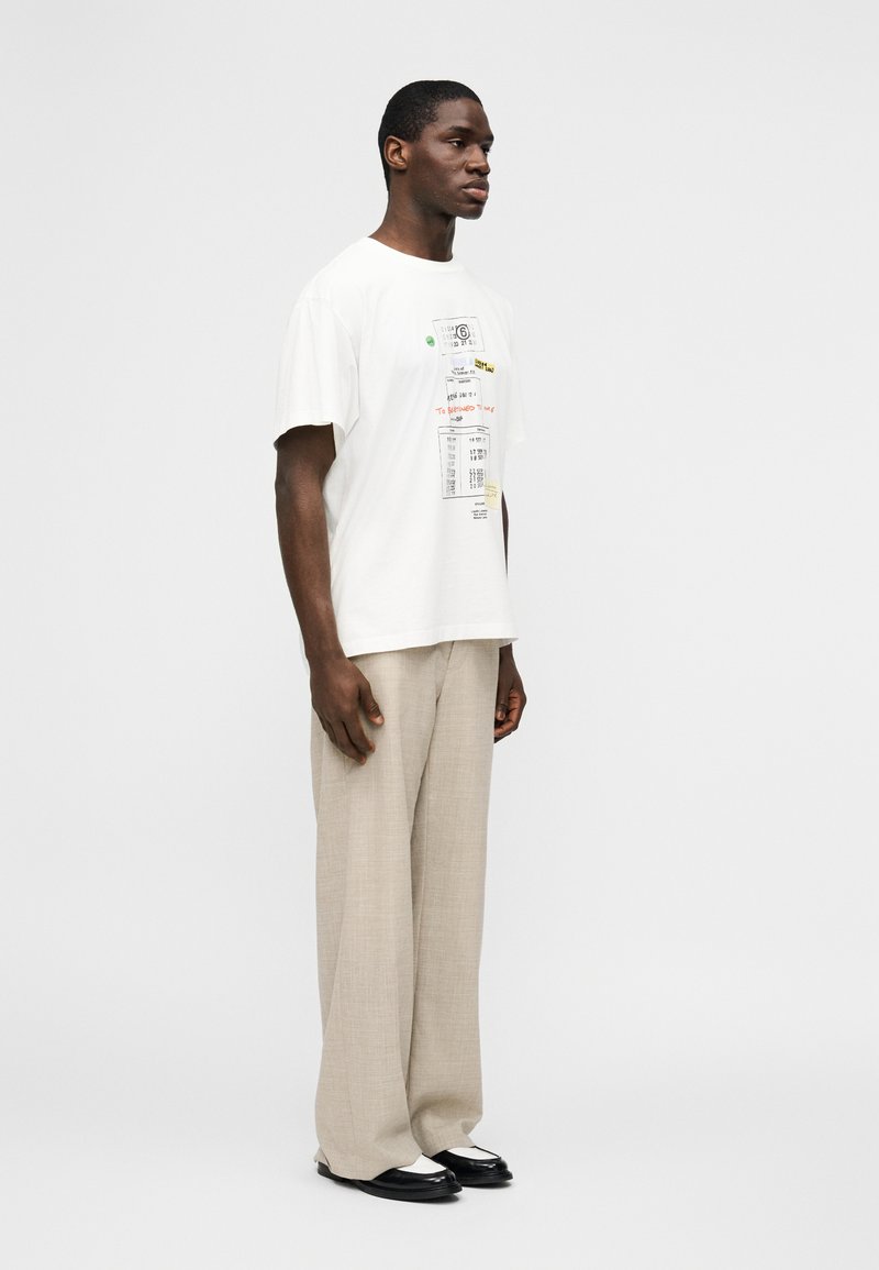 Uomo in piedi che indossa una t-shirt bianca con stampa grafica, pantaloni beige a gamba larga e scarpe bianche e nere su uno sfondo bianco uniforme.