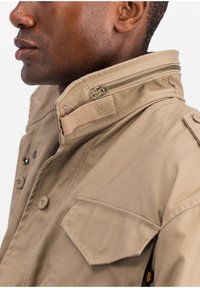 Khaki jacket with a high collar, zipper detail, button closures, and a front pocket. Made of a durable fabric with a smooth texture.