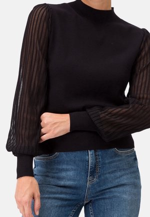 Top noir ajusté avec des manches longues ballon transparentes rayées, associé à un jean bleu, montrant le torse et les bras du modèle.